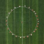 Aerial view of a soccer team gathered in a huddle, forming a circle around one player at the centre of the pitch.