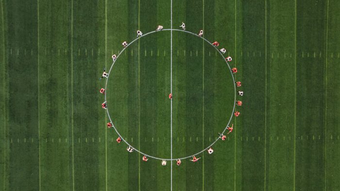 Aerial view of a soccer team gathered in a huddle, forming a circle around one player at the centre of the pitch.
